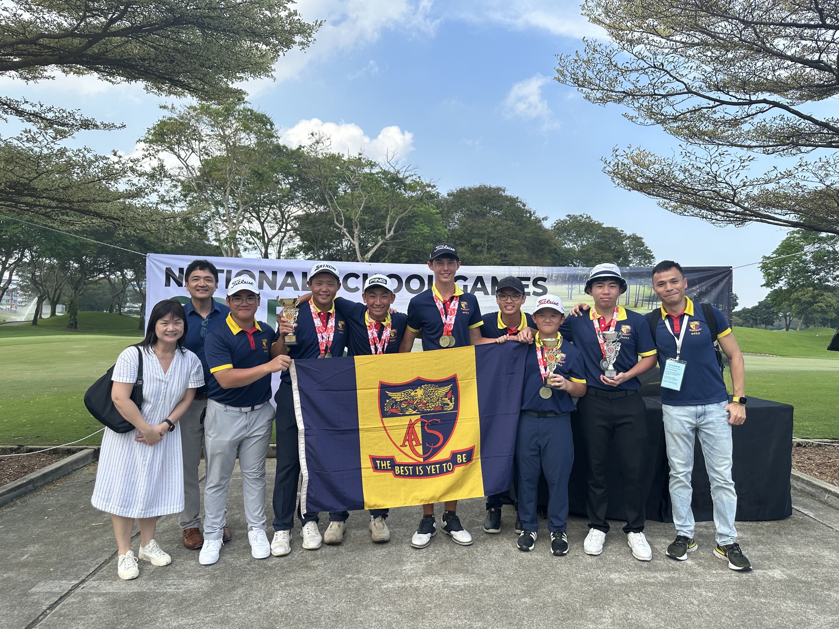 A group of young golfers with medals and trophies hold an ACS school flag at the National School Games golf event.